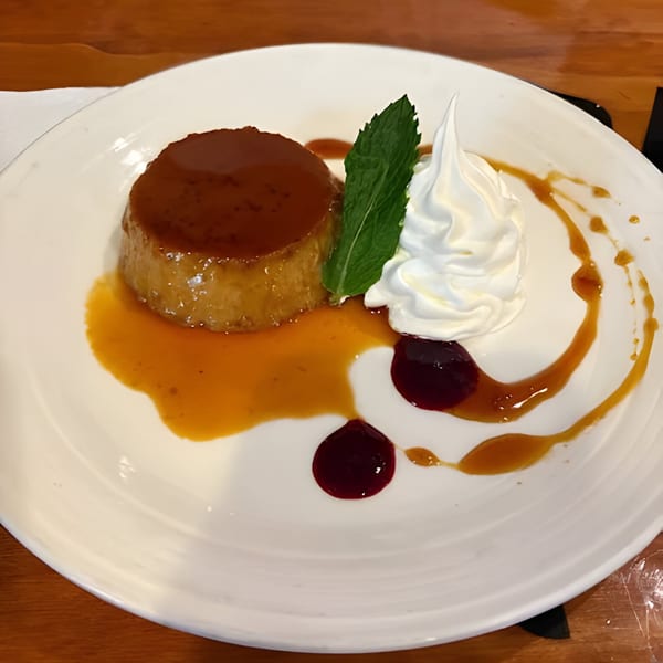 Flan dessert with caramel sauce, whipped cream, mint leaf, and berry compote on a white plate.