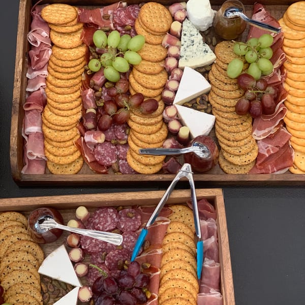 Two wooden trays overflowing with crackers, cheeses, grapes, cured meats, and jams.