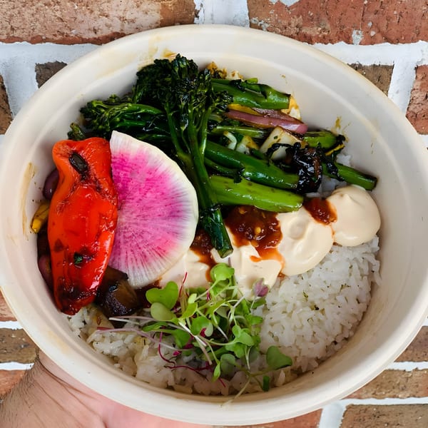 A vibrant rice bowl with grilled red pepper, broccolini, watermelon radish, and sauces, held against a brick wall.