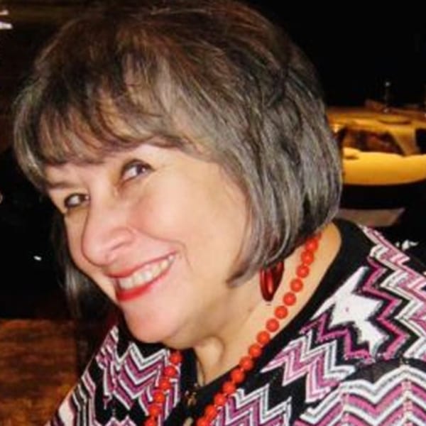 A smiling woman with short brown hair, red lipstick, and a red beaded necklace.