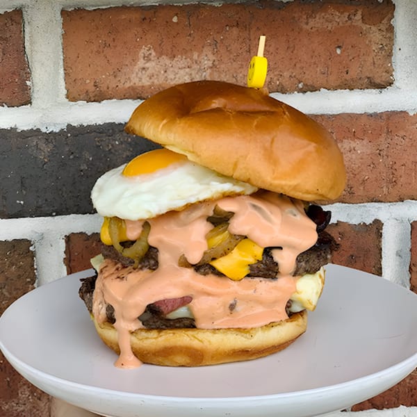 A double patty burger with cheese, fried egg, grilled onions, and pink sauce on a white plate.