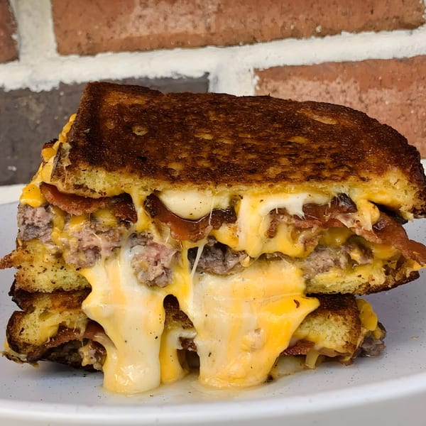 A gooey, cheesy burger melt with bacon and ground beef on toasted bread, dripping cheese onto a white plate.