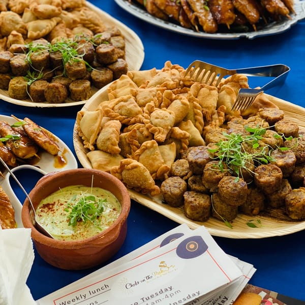 Platters of empanadas, meat skewers, and round appetizers with a green dip, on a blue tablecloth.