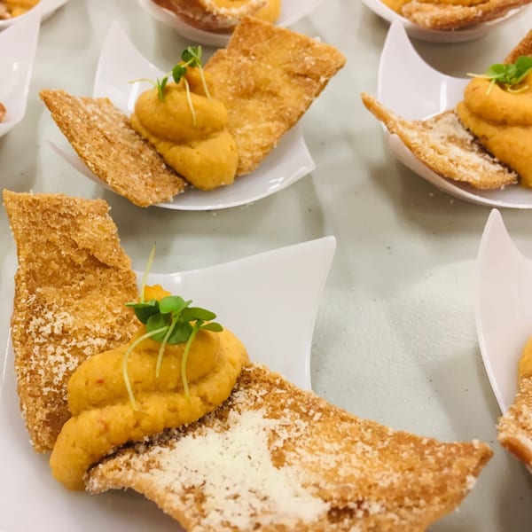 Close-up of several appetizers with crispy flatbread, orange dip, and green garnish on white dishes.