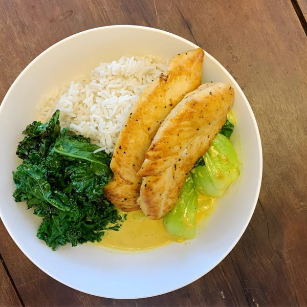 Grilled chicken, white rice, kale, and bok choy in a yellow sauce, served in a white bowl.
