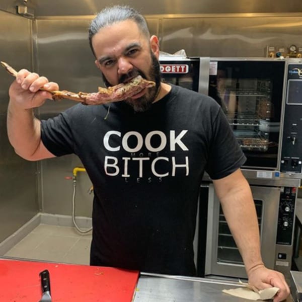A chef in a black t-shirt with "COOK MORE BITCH LESS" printed on it, eating a large piece of meat off a bone in a kitchen.